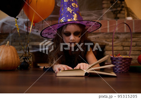Adorable Caucasian child girl looking like a witch in a wizard hat, lying on the floor and studying a spell book, surrounded by a fireplace covered with cobwebs, a pumpkin, black and orange balloons Adorable Caucasian child girl looking like a witch in a wizard hat, lying on the floor and studying a spell book, surrounded by a fireplace covered with cobwebs, a pumpkin, black and orange balloons 95315205