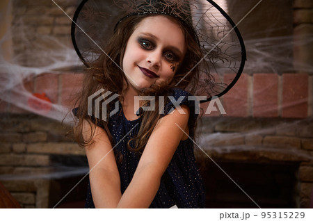 Caucasian cute baby girl with a black spider drawn on her cheek, wearing a wizard's hat, a little sorceress, smiles sweetly at camera, posing against a spider web covered fireplace and Halloween Jack Caucasian cute baby girl with a black spider drawn on her cheek, wearing a wizard's hat, a little sorceress, smiles sweetly at camera, posing against a spider web covered fireplace and Halloween Jack 95315229