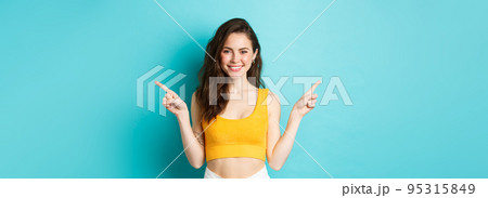 Choose your turn. Smiling beautiful woman with perfect body in crop top, pointing fingers sideways, showing two ways, standing over blue background Choose your turn. Smiling beautiful woman with perfect body in crop top, pointing fingers sideways, showing two ways, standing over blue background 95315849