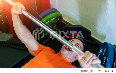 Amazing image of a young man doing bench press with a maximum weight. He screams in the effort. Exciting and motivational photo. Amazing image of a young man doing bench press with a maximum weight. He screams in the effort. Exciting and motivational photo. 95316313