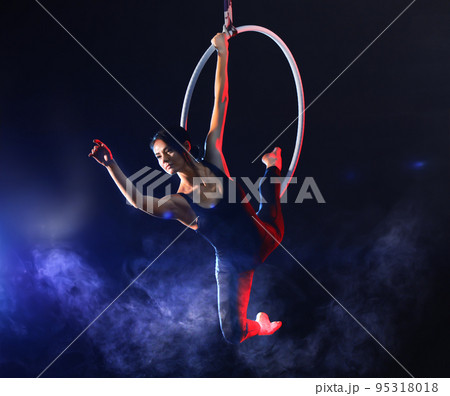 Young woman performing acrobatic element on aerial ring against dark background 95318018