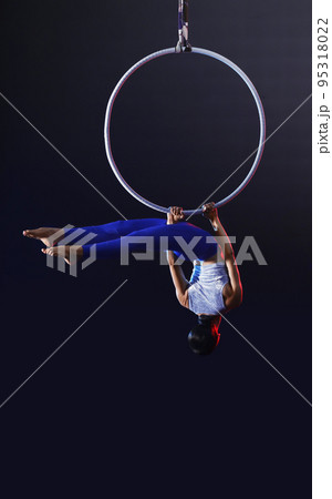 Young woman performing acrobatic element on aerial ring against dark background Young woman performing acrobatic element on aerial ring against dark background 95318022