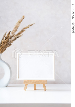 Empty picture frame mock up in minimalist interior with dried plant in vase on white table. Empty picture frame mock up in minimalist interior with dried plant in vase on white table. 95323384
