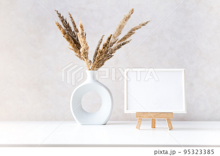 Empty picture frame mock up in minimalist interior with dried plant in vase on white table. 95323385