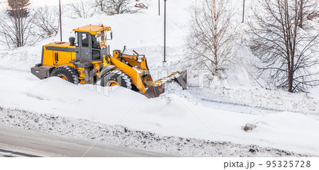 Snow clearing. Tractor clears the way after heavy snowfall. A large orange tractor removes snow from the road and clears the sidewalk. Cleaning roads in the city from snow in winter. 95325728