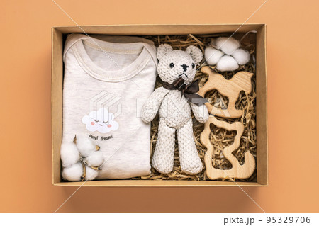 Gift basket with gender neutral baby garment and accessories. Care box of organic newborn cotton clothes, teddy, cotton in box on orange background Flat lay Top view. Gift basket with gender neutral baby garment and accessories. Care box of organic newborn cotton clothes, teddy, cotton in box on orange background Flat lay Top view. 95329706