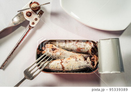 Open can of sardines in oil next to a can opener, a plate and a fork on a white marble-like surface. 95339389