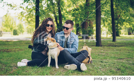 Excited young people loving couple are fussing cute dog shiba inu scratching its fur, talking and laughing sitting on grass in park. People and domestic animals concept. Excited young people loving couple are fussing cute dog shiba inu scratching its fur, talking and laughing sitting on grass in park. People and domestic animals concept. 95340610