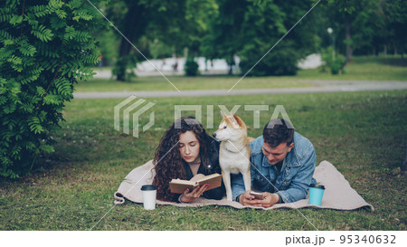 Young family spending weekend in the park, woman reading book, man using smartphone, cute dog resting between them. Guy is showing phone screen to his laughing wife. Young family spending weekend in the park, woman reading book, man using smartphone, cute dog resting between them. Guy is showing phone screen to his laughing wife. 95340632