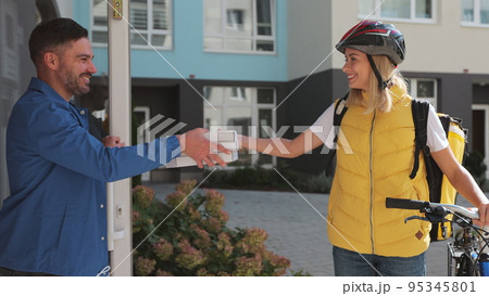 Close up of handsome man open door to a pizza delivery woman wearing yellow thermal backpack on a bike and recieves cardboard boxes of pizza. Food delivery concept. Close up of handsome man open door to a pizza delivery woman wearing yellow thermal backpack on a bike and recieves cardboard boxes of pizza. Food delivery concept. 95345801