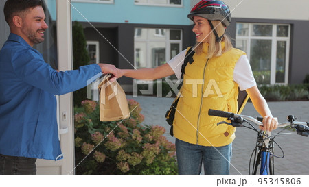 Close up man customer open door to a food delivery woman wearing yellow thermal backpack on a bike and recieves order or package with food. Food delivery concept. Close up man customer open door to a food delivery woman wearing yellow thermal backpack on a bike and recieves order or package with food. Food delivery concept. 95345806