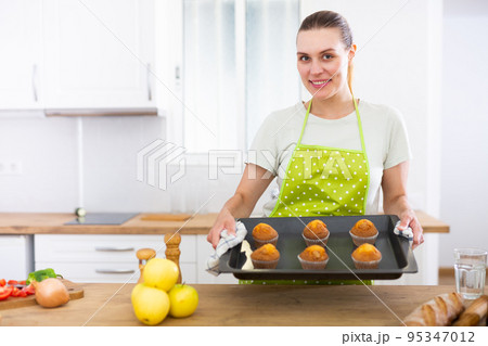 Cheerful housewife standing in kitchen with baking tray of muffins Cheerful housewife standing in kitchen with baking tray of muffins 95347012