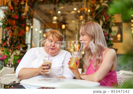 Young daughter and her mother in wheelchair sitting at the table in cafe with drinks and having fun Young daughter and her mother in wheelchair sitting at the table in cafe with drinks and having fun 95347142