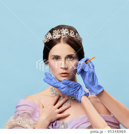 Half-length portrait of beautiful young woman in vintage dress and tiara as royal person makes beauty injections on blue background. 95348969