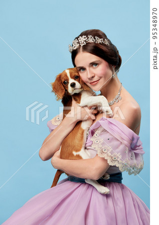 Beloved pet. Portrait of young charming woman in lilac color vintage dress as medieval princess holding small King Charles spaniel dog on blue background. 95348970