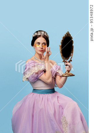 Face skin care. Beautiful charming woman in lilac color vintage dress as a royal person, princess looking in mirror at herself on blue background. 95348972