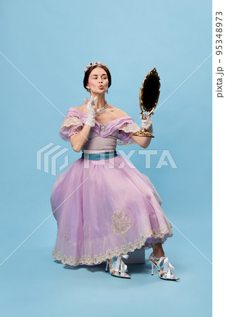 Self loving. Beautiful charming woman in lilac color vintage dress as a royal person, princess looking in mirror at herself on blue background. 95348973