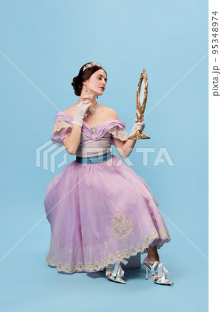 Young and pretty. Beautiful charming woman in lilac color vintage dress as a royal person, princess looking in mirror at herself on blue background. 95348974