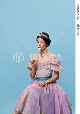 Flirt. Portrait of sensual young girl in lilac color medieval dress as young queen or princess tasting lollipop. Eras comparison, beauty, art, emotions and vintage fashion style 95348985