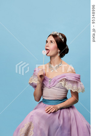 Sweets. Sensual girl in lilac color medieval dress as young queen or princess tasting lollipop. Eras comparison, beauty, art, emotions and vintage fashion style 95348996