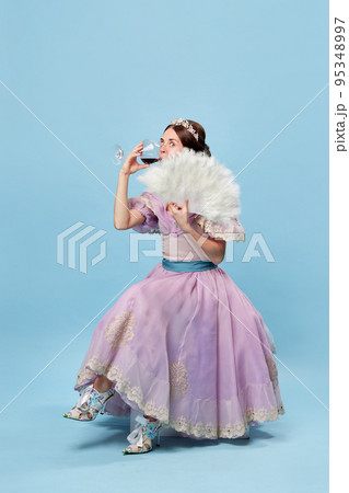 Tasting wine. Cute young girl in lilac color medieval dress as young queen or princess on blue background. Eras comparison, beauty, art, emotions and vintage fashion style 95348997