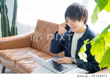 パソコンとスマホで作業する20代男性-リビングのソファーと観葉植物 パソコンとスマホで作業する20代男性-リビングのソファーと観葉植物 95349971