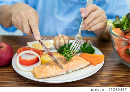 Asian senior or elderly old lady woman patient eating Salmon steak breakfast with vegetable healthy food while sitting and hungry on bed in hospital. 95350447