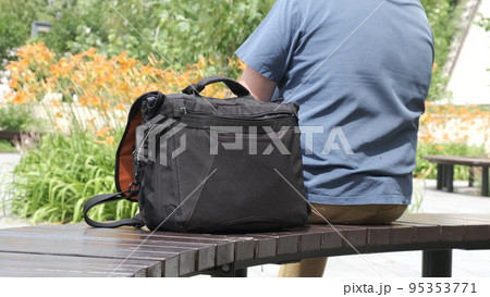Bag is on park bench next to its owner. Man is resting with photo bag next to him. 95353771