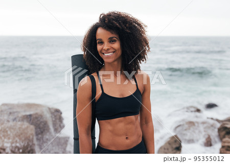 Portrait of a muscular woman with a mat standing by the ocean and looking away Portrait of a muscular woman with a mat standing by the ocean and looking away 95355012