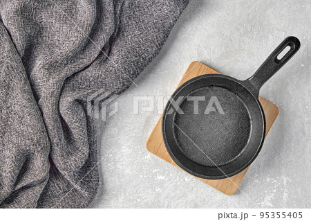 Black cast iron empty frying pan for one person on a wooden stand and a gray kitchen towel on a gray concrete background 95355405