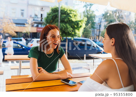 Two pretty girlfriends talking while sitting in a bar outdoors Two pretty girlfriends talking while sitting in a bar outdoors 95355800