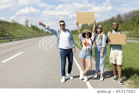 Happy diverse friends standing on highway with empty signs, stopping car, going on trip together, outdoors 95357002