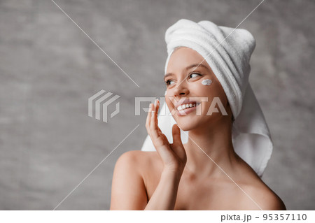 Female skincare concept. Lady applying moisturizer cream under eyes, standing in bathroom, looking aside and smiling Female skincare concept. Lady applying moisturizer cream under eyes, standing in bathroom, looking aside and smiling 95357110