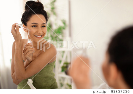 Attractive young brunette woman applying moisturizing serum onto her face near mirror at home, free space Attractive young brunette woman applying moisturizing serum onto her face near mirror at home, free space 95357256