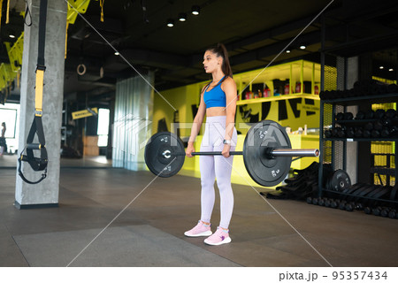 Strong young woman doing sit-ups with a barbell 95357434