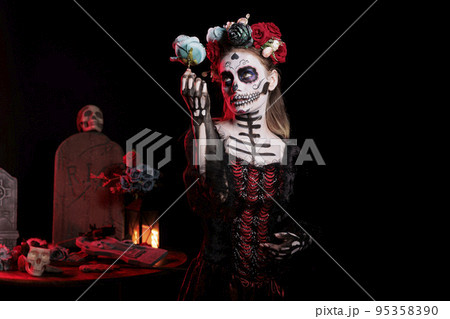 Spooky entity of death looking like santa muerte in studio, posing with roses and wearing glamour costume. Female goddess acting horror and celebrating mexican dios de los muertos holiday. 95358390