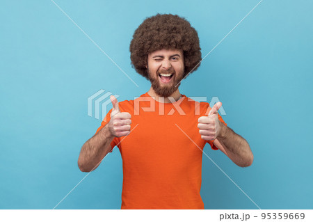 Portrait of man with Afro hairstyle wearing orange T-shirt showing thumbs up, winking and smiling excitedly, enjoying excellent result. Indoor studio shot isolated on blue background. 95359669