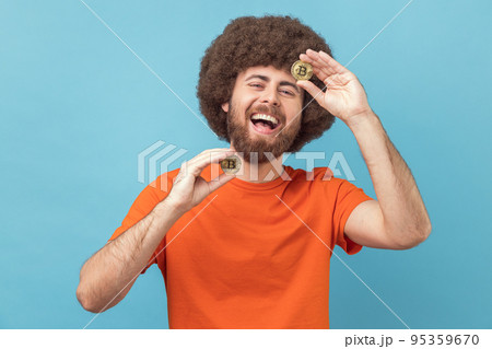 Portrait of funny laughing man with Afro hairstyle wearing orange T-shirt standing holding bitcoins, paying attention at new digital cryptocurrency. Indoor studio shot isolated on blue background. Portrait of funny laughing man with Afro hairstyle wearing orange T-shirt standing holding bitcoins, paying attention at new digital cryptocurrency. Indoor studio shot isolated on blue background. 95359670
