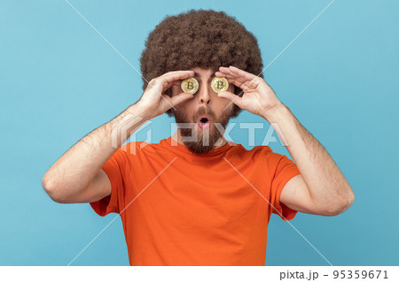 Portrait of astonished man with Afro hairstyle wearing orange T-shirt covering eyes with golden bitcoin, looking at camera through btc coin. Indoor studio shot isolated on blue background. Portrait of astonished man with Afro hairstyle wearing orange T-shirt covering eyes with golden bitcoin, looking at camera through btc coin. Indoor studio shot isolated on blue background. 95359671