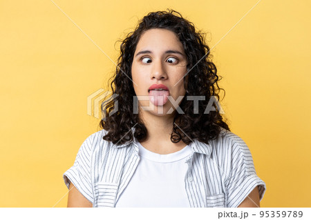Portrait of woman with dark wavy hair standing with crossed eyes and tongue out, having foolish facial expression, childish behavior. Indoor studio shot isolated on yellow background. 95359789