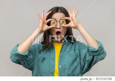 Portrait of amazed surprised woman covering eyes with golden bitcoin, looking camera through btc coin, keeps mouth open, wearing casual style jacket. Indoor studio shot isolated on gray background. 95359790