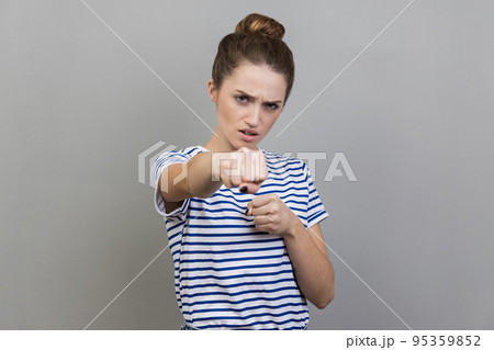 Portrait of aggressive woman wearing striped T-shirt standing holding clenched fists up ready to boxing, fighting spirit, boxing. Indoor studio shot isolated on gray background. 95359852