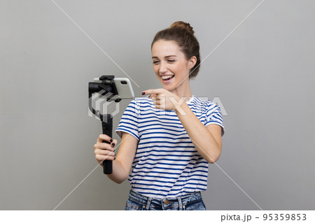 Portrait of attractive woman wearing striped T-shirt pointing to cell phone camera with smile, streaming, talking with followers, uses steadicam. Indoor studio shot isolated on gray background. 95359853