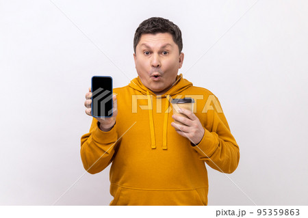 Portrait of astonished surprised man of middle age holding smart phone with blank screen and disposable cup of coffee, wearing urban style hoodie. Indoor studio shot isolated on white background. Portrait of astonished surprised man of middle age holding smart phone with blank screen and disposable cup of coffee, wearing urban style hoodie. Indoor studio shot isolated on white background. 95359863
