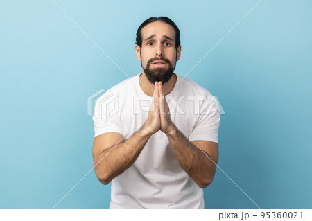 Please, I'm begging forgive. Portrait of upset worried man with beard wearing white T-shirt looking with imploring desperate grimace, praying for help. Indoor studio shot isolated on blue background. Please, I'm begging forgive. Portrait of upset worried man with beard wearing white T-shirt looking with imploring desperate grimace, praying for help. Indoor studio shot isolated on blue background. 95360021