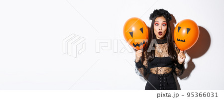 Image of surprised asian woman in witch costume celebrating halloween, holding balloons with scary faces, standing over white background Image of surprised asian woman in witch costume celebrating halloween, holding balloons with scary faces, standing over white background 95366031
