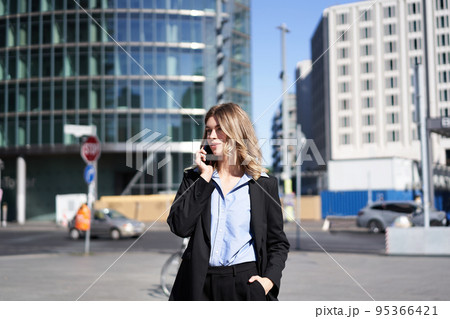 Portrait of businesswoman in suit, standing on street with mobile phone, on call with someone, smiling while having telephone conversation Portrait of businesswoman in suit, standing on street with mobile phone, on call with someone, smiling while having telephone conversation 95366421