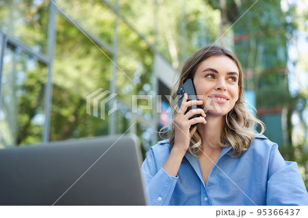 Portrait of young businesswoman sitting outside in park, talking on mobile phone, using laptop. Young woman, student working outdoors, calling, talking on telephone Portrait of young businesswoman sitting outside in park, talking on mobile phone, using laptop. Young woman, student working outdoors, calling, talking on telephone 95366437