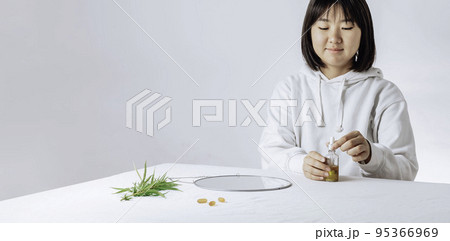 Asian woman with cbd liquid oil made from cannabis extract for a natural skin treatment. Portrait of young woman with cannabis and mirror at the table. Cosmetology and treatment. 95366969