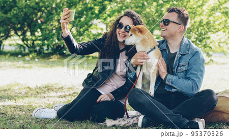 Happy couple is taking selfie with their dog using smartphone sitting on grass in the park, people are posing, talking and petting shiba inu puppy. Photograph and animals concept. 95370326